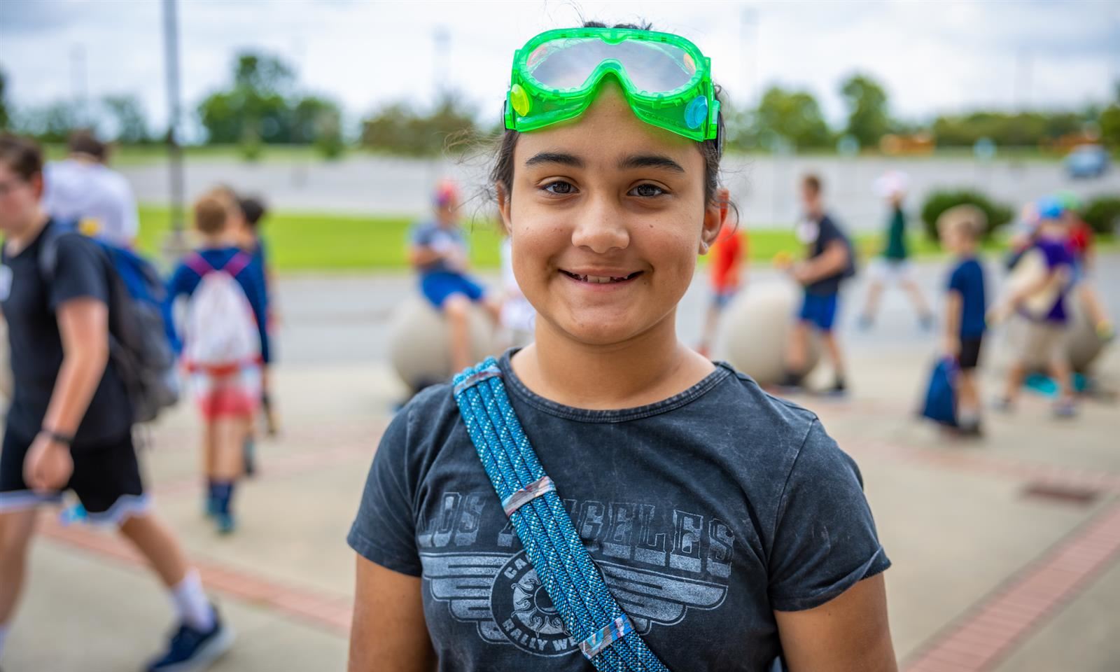 A girl wearing a dark t-shirt and a pair of green lab goggles on her head smiles